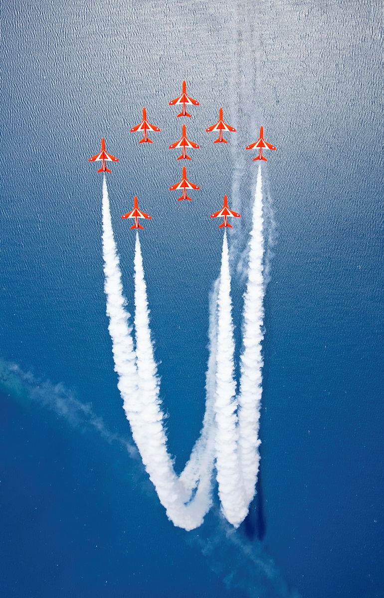 Aerial view of the Red Arrows flying in formation over the sea, with white slip smoke.